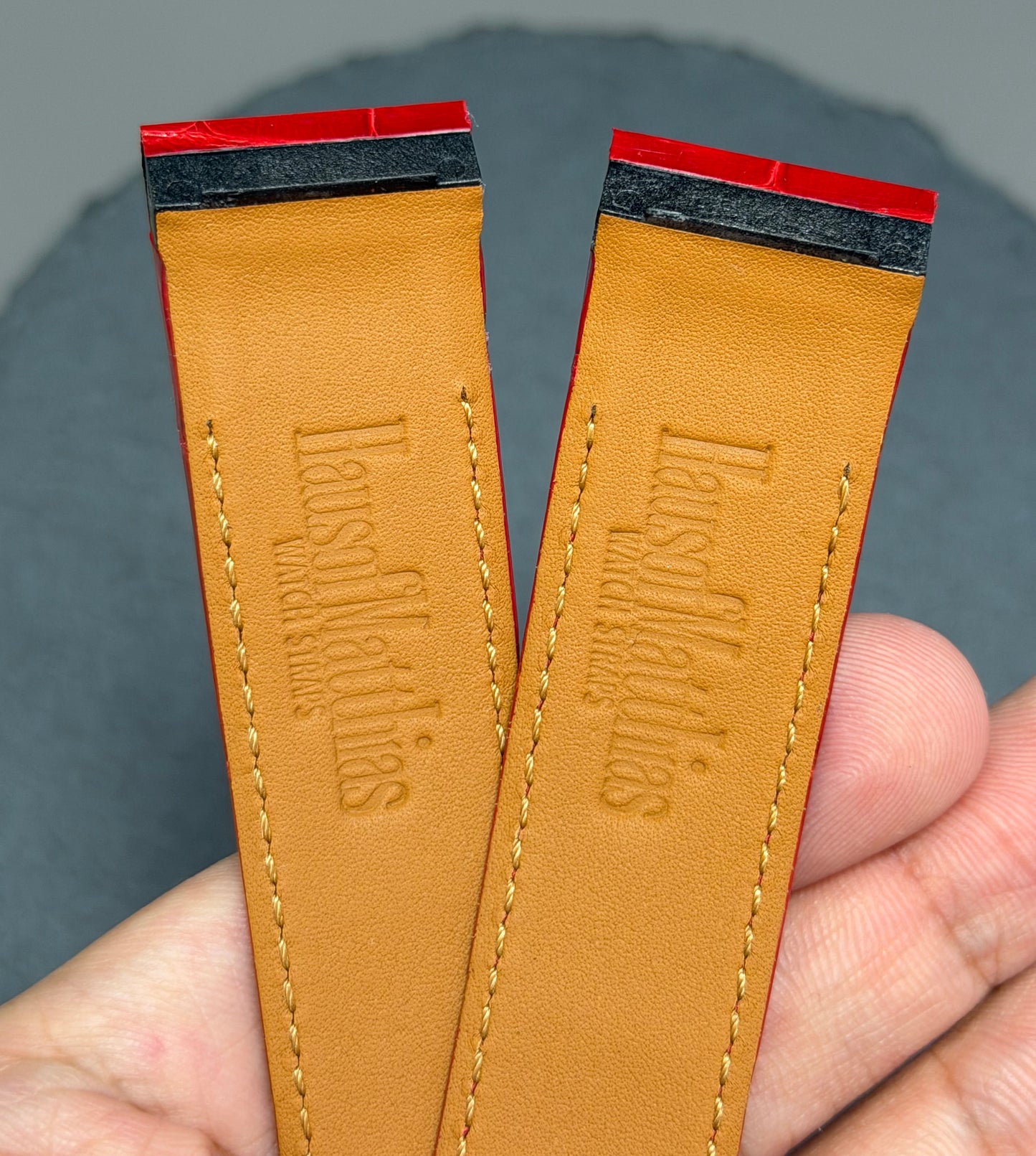 Two brown leather straps with 'HausofMathias' branding held by a hand against a gray background.