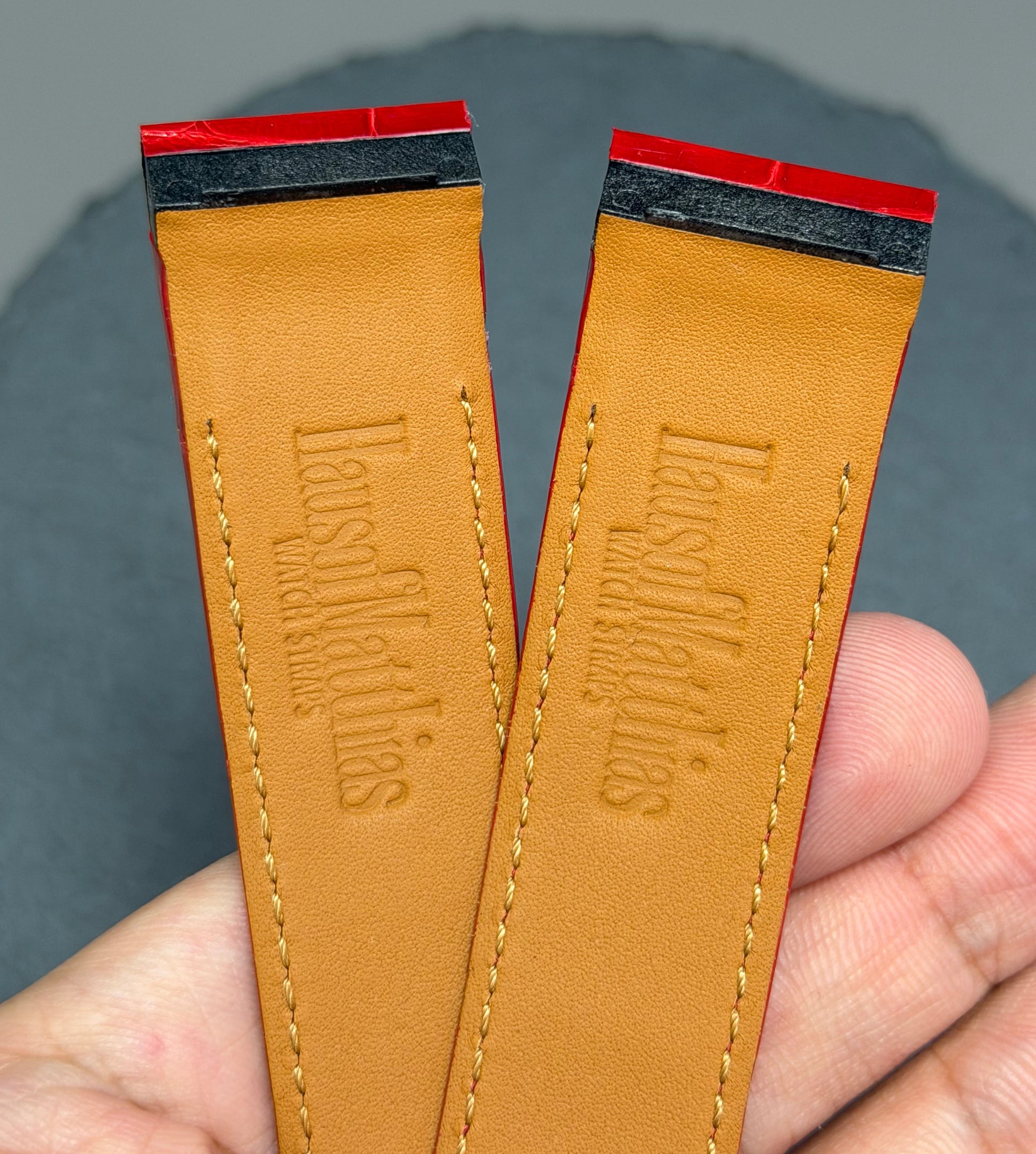 Two brown leather straps with 'HausofMathias' branding held by a hand against a gray background.
